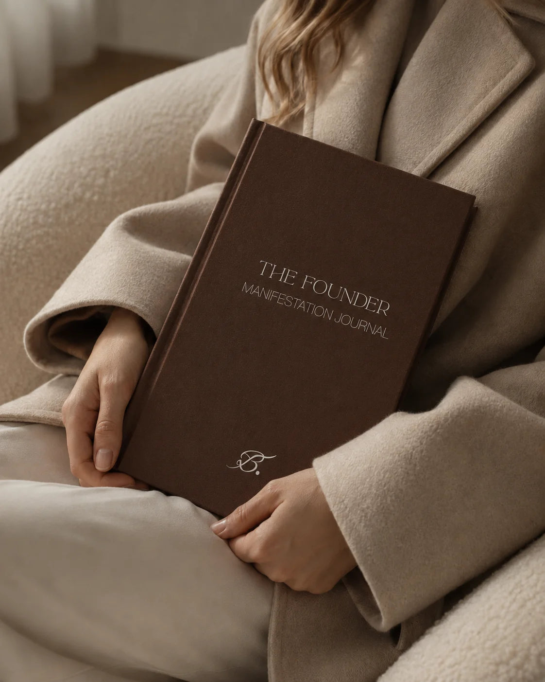 The Founder Manifestation Journal by Bianca Da Silva, a dark brown hardcover journal held against a beige coat