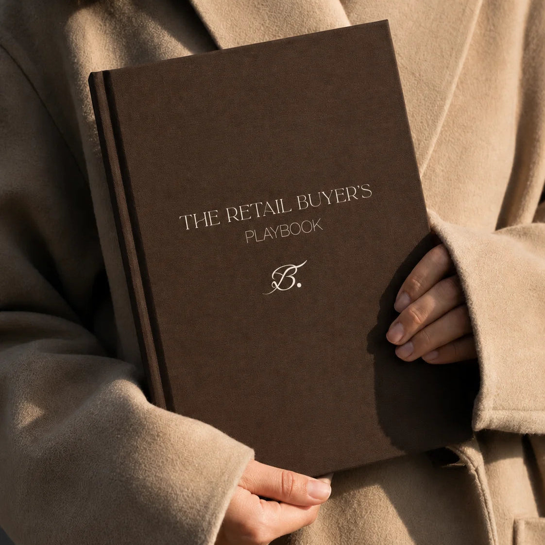 The Retail Buyer's Playbook by Bianca Da Silva, a dark brown hardcover book held against a beige coat