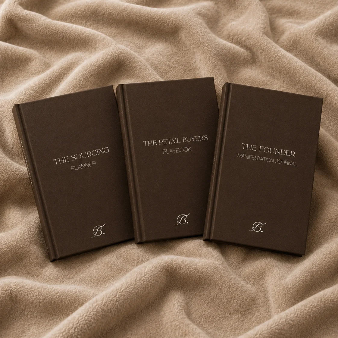 The Sourcing Planner, The Retail Buyer's Playbook and The Founder Manifestation Journal by Bianca Da Silva, dark brown hardcover journals with gold monogram on a beige blanket