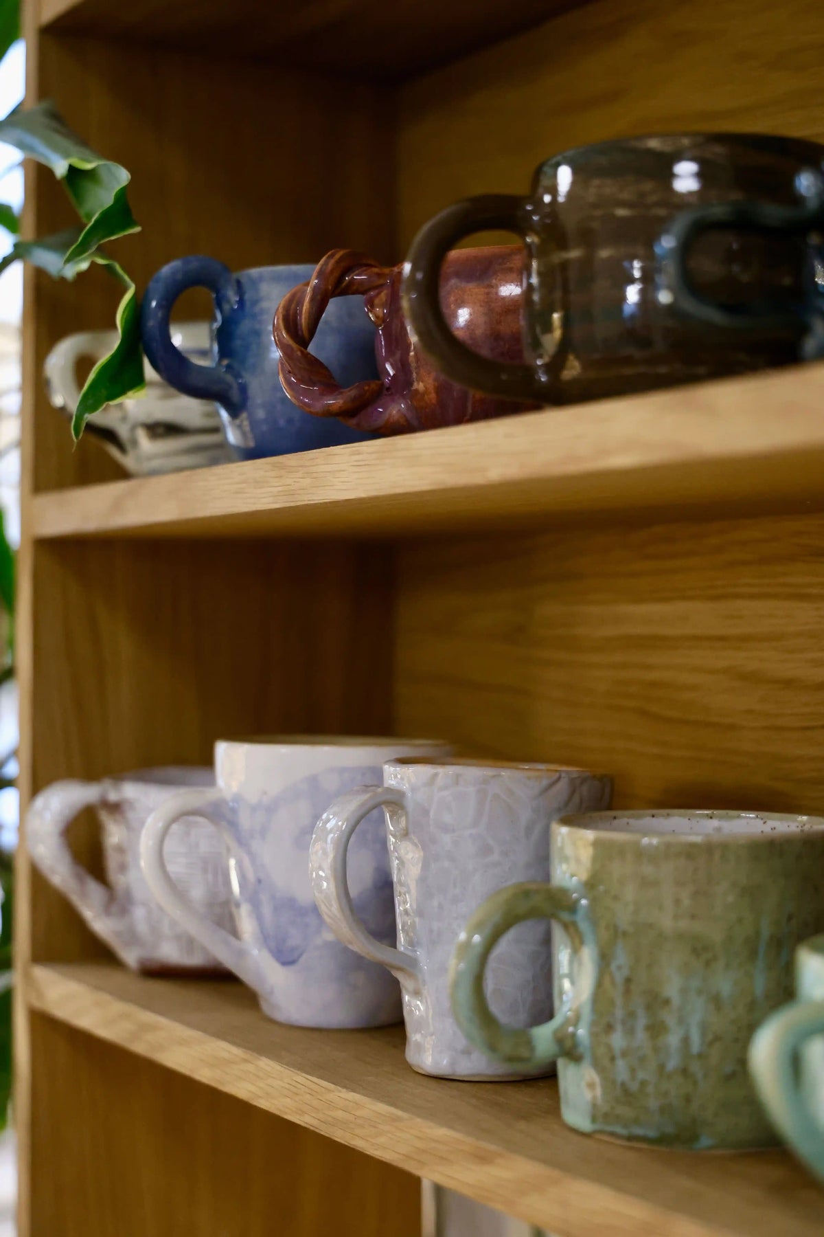 Collection of handmade ceramic mugs in various colours and glazes displayed on a wooden shelf